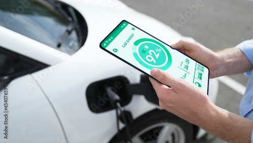Businessman holding smartphone display battery status interface by smart EV mobile application while EV car recharging electricity from charging station in car park. Peruse
