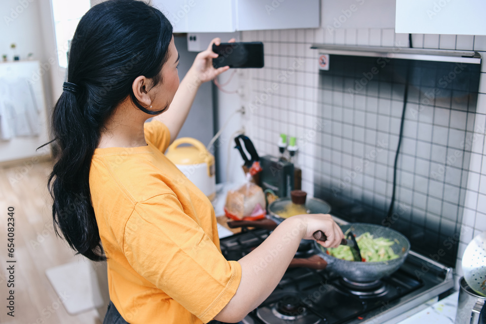 Young Asian woman taking selfie with smartphone while cooking in the kitchen - obrazy, fototapety, plakaty 