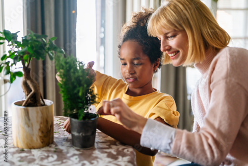 Happy caucasian mother and her adopted african american daughter taking care of plants indoors.