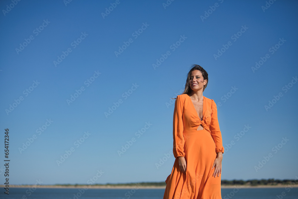 Beautiful young woman in orange dress, posing, happy and smiling, on the beach, with the wind ruffling her hair and dress. Concept beauty, fashion, trend, air, beach. Beautiful young woman in orange dress, posing, happy and smiling, on the beach, with the wind ruffling her hair and dress. Concept beauty, fashion, trend, air, beach.