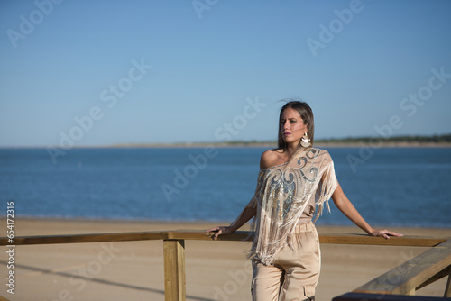 Young and beautiful woman with sequined and fringed shirt, leaning on a wooden railing, looking at infinity by the sea. Concept beauty, fashion, trend, empowerment.