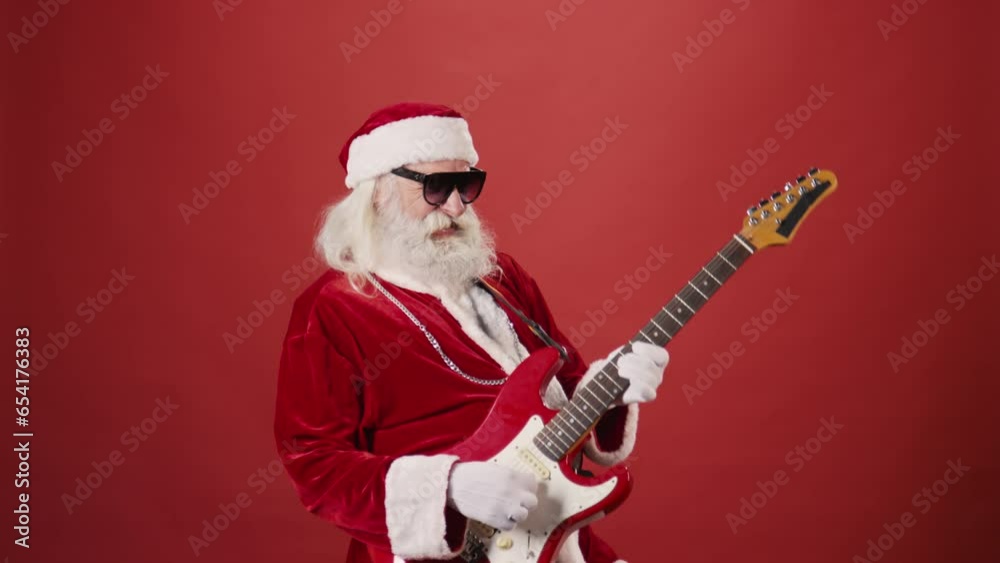 Medium shot of bearded elderly man in Santa Claus costume, with sunglasses and silver chain, energetically playing electric guitar, on plain red background. Template, copy space Medium shot of bearded elderly man in Santa Claus costume, with sunglasses and silver chain, energetically playing electric guitar, on plain red background. Template, copy space