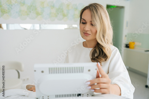 Portrait of professional female beautician typing on screen of device in beauty salon during aesthetic procedure. Concept of non-surgery cosmetology, professional skincare, beauty treatment