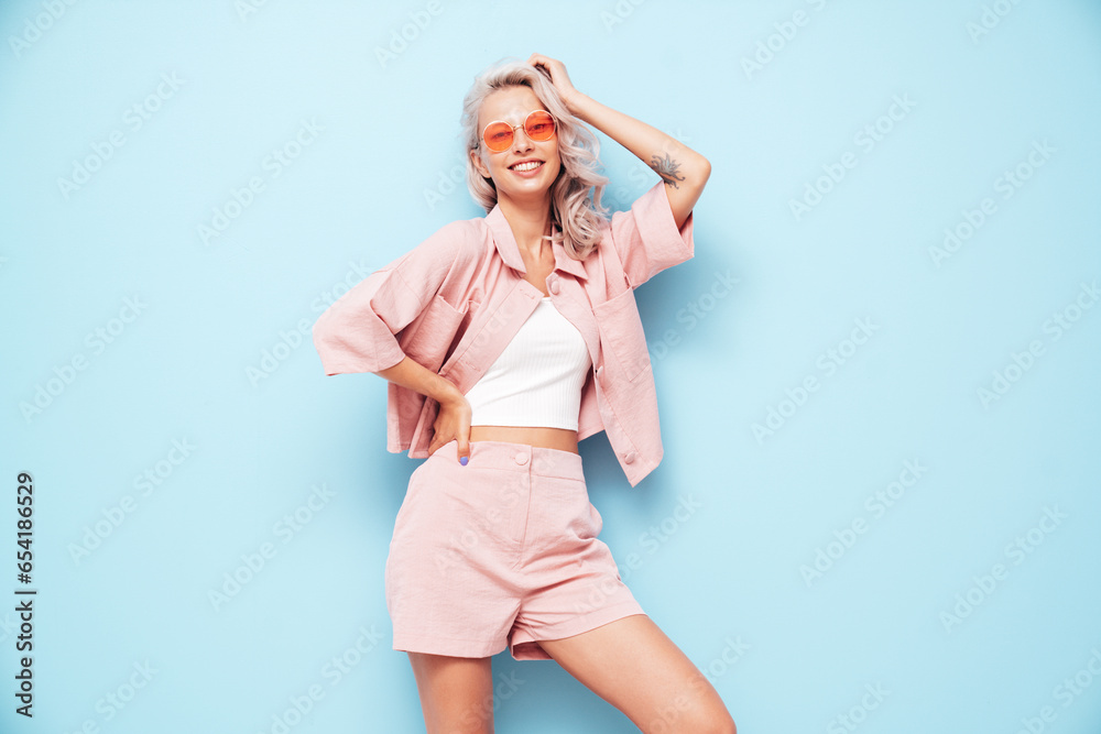 Young beautiful smiling female in trendy summer pink shorts and t-shirt. Carefree woman posing near blue wall in studio. Positive blond model having fun. Cheerful and happy. in sunglasses Young beautiful smiling female in trendy summer pink shorts and t-shirt. Carefree woman posing near blue wall in studio. Positive blond model having fun. Cheerful and happy. in sunglasses