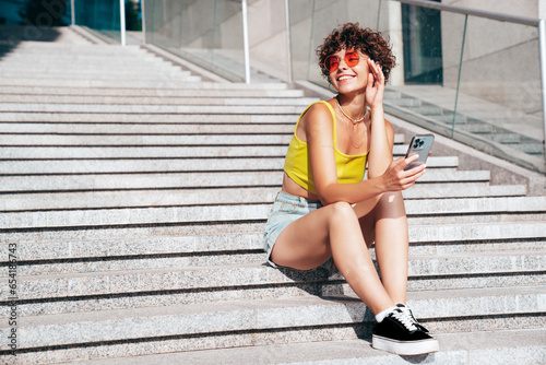 Young beautiful smiling hipster woman in trendy summer clothes. Carefree woman with curls hairstyle, in the street. Positive model holds mobile phone, looks at cellphone screen, uses smartphone apps