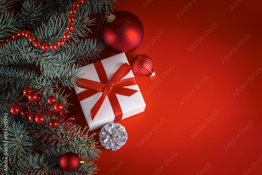 White box with a gift tied with a red ribbon, fir branches and Christmas decorations on a red background White box with a gift tied with a red ribbon, fir branches and Christmas decorations on a red background