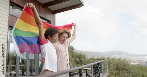 Happy diverse gay male couple holding rainbow lgbt flag on balcony at home, slow motion