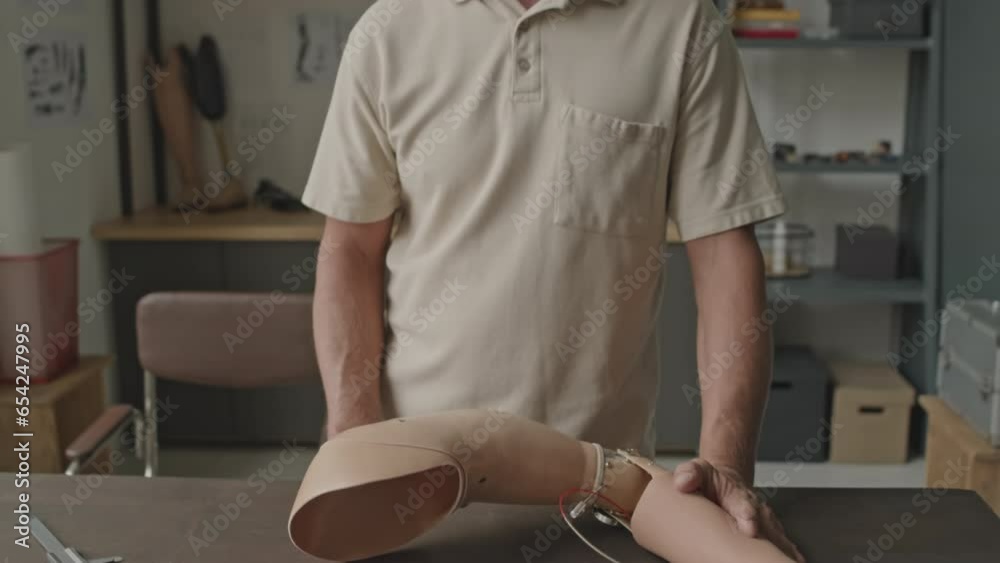 Tilt up portrait of mature Caucasian specialist with arm prosthesis looking at camera while standing by work table in small workshop