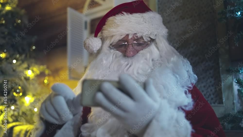 Santa Claus is sitting in a chair in front of the fireplace at night and using smartphone, scrolling and surfing on the internet. Christmas as a holiday concept. Santa Claus is sitting in a chair in front of the fireplace at night and using smartphone, scrolling and surfing on the internet. Christmas as a holiday concept.