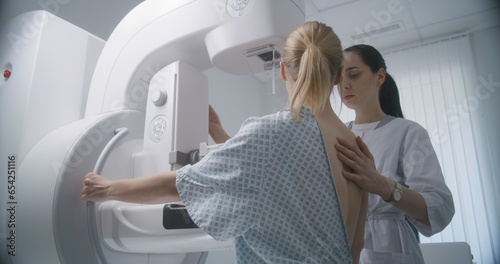 Hospital radiology room. Caucasian woman stands during mammography screening examination in clinic. Female doctor adjusts modern mammogram machine for patient, uses computer. Breast cancer prevention. photo