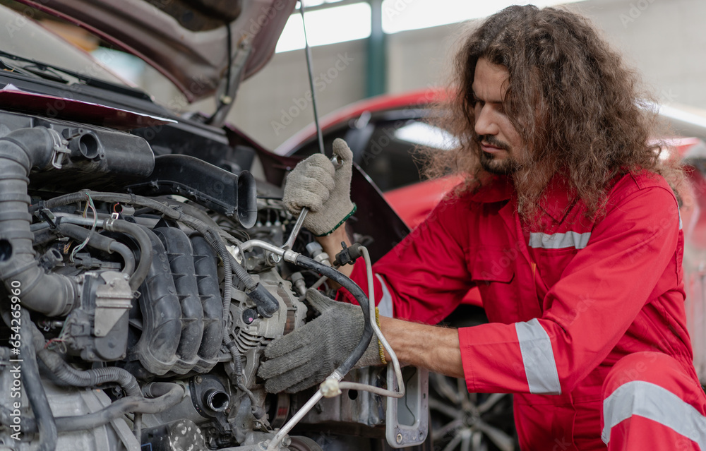Auto mechanic working in vehicle repair service garage. Handsome long hair repairman inspecting car engine problem, damage motor, fix or change part. People in automobile maintenance business concept