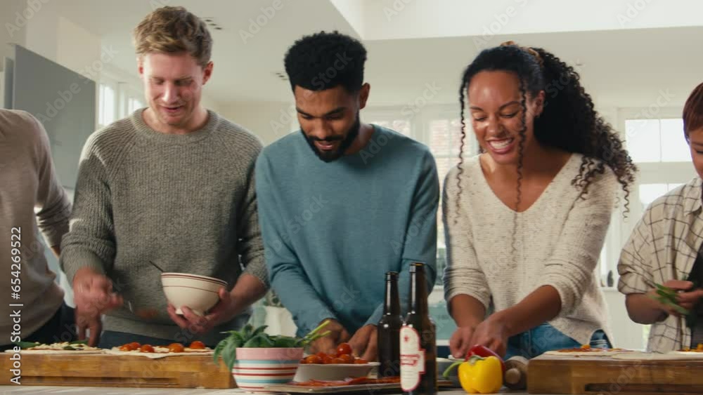 Group of friends at home in kitchen adding toppings to homemade pizzas for party together 