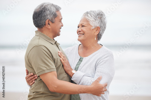 Elderly, couple and hug with smile on beach in nature together for holiday, relax and connection outdoor. Commitment, man or woman with love embrace for peace, trust and vacation at ocean or sea
