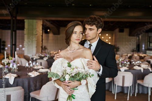 elegant groom hugging young and charming bride with wedding bouquet in modern banquet hall