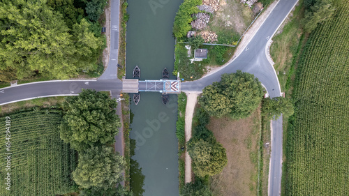 Experience the enchanting beauty of nature from above with this top-down aerial view. Roads converge toward a charming drawbridge that spans a serene canal meandering through lush forests and fertile