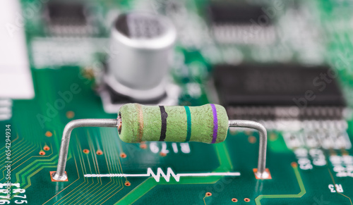 Closeup of resistor and white electronic symbol on green PCB surface. Small component with color code and two metal wire terminals on printed circuit board and blurry capacitor or chips in background. photo