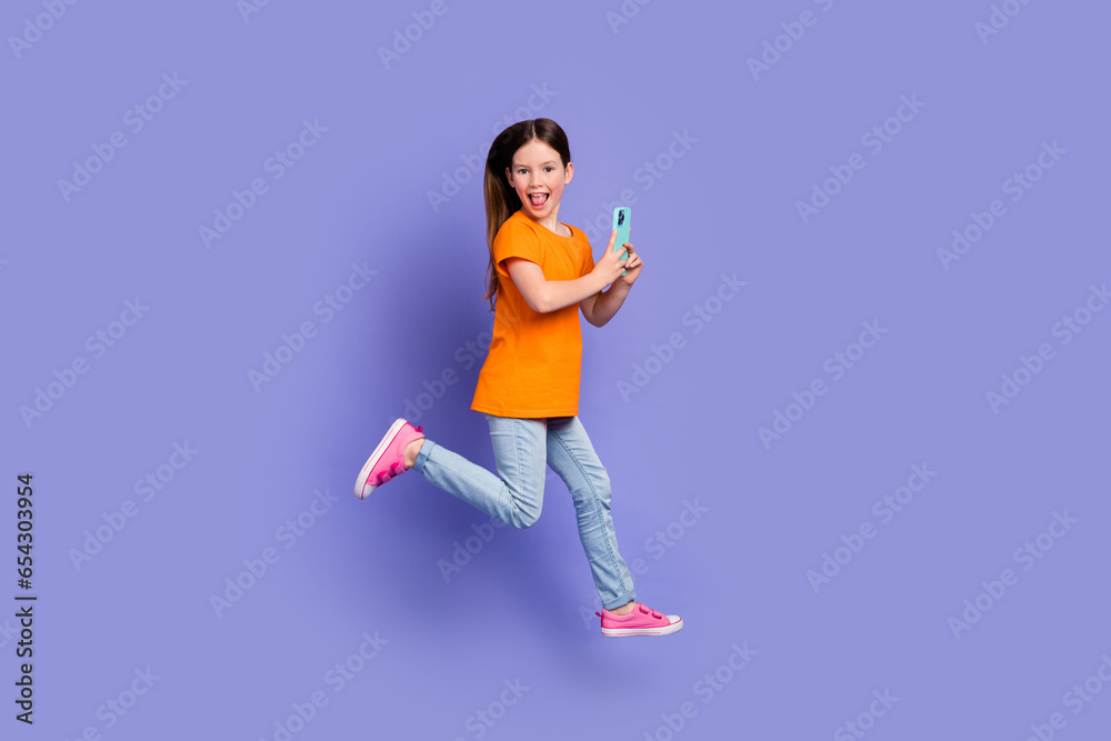 Full size photo of positive schoolgirl wear stylish t-shirt hold smartphone run in empty space isolated on violet color background
