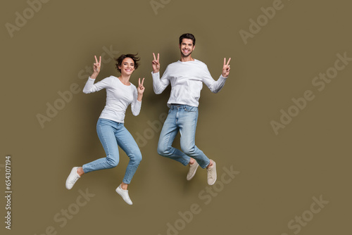 Full length photo of pretty charming married couple wear white shirts jumping showing v-signs empty space isolated brown color background