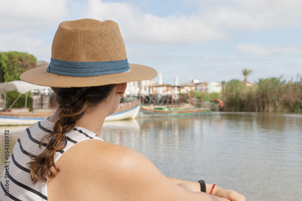 A young woman, with her back to the camera and wearing a hat, enjoys the picturesque view of the calm waters of a lakeside harbor