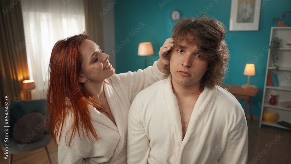 A close up of a young couple is depicted. The man with a disgruntled, face looks at the camera as if in a mirror. The girl joyfully with a smile on her face at this time corrects and styles his hair A close up of a young couple is depicted. The man with a disgruntled, face looks at the camera as if in a mirror. The girl joyfully with a smile on her face at this time corrects and styles his hair