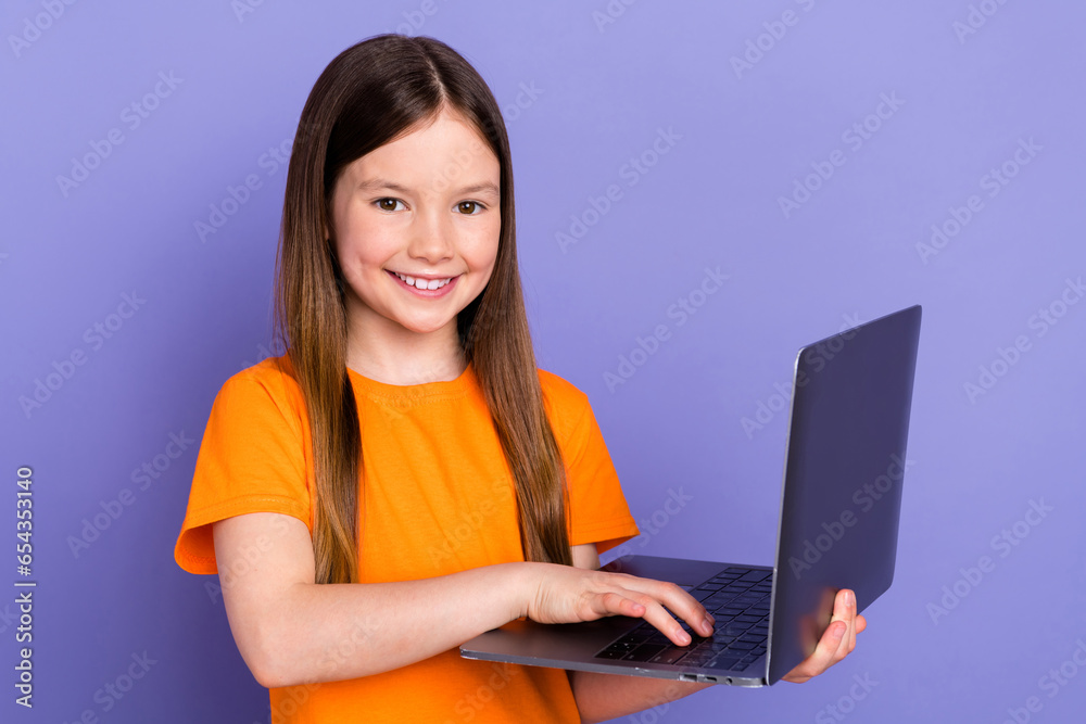 Photo of optimistic smile young schoolgirl wear orange t shirt holding netbook communicating remote isolated on violet color background Photo of optimistic smile young schoolgirl wear orange t shirt holding netbook communicating remote isolated on violet color background