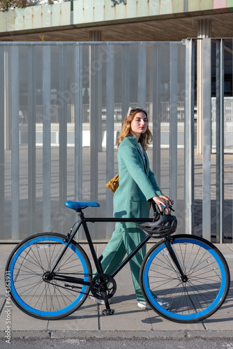 Stylish red-haired woman in green suit walking with bicycle at the financial district with modern building on the background