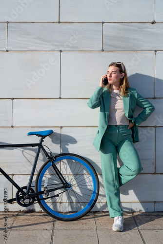 Young empowered ginger-haired woman in green suit talking on smartphone standing with bicycle at marble background