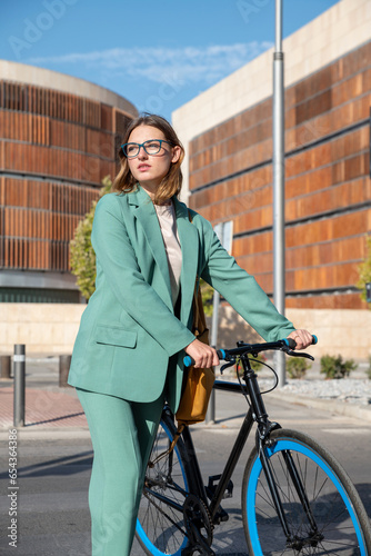 Stylish red-haired woman in green suit walking with bicycle at the financial district with modern building on the background