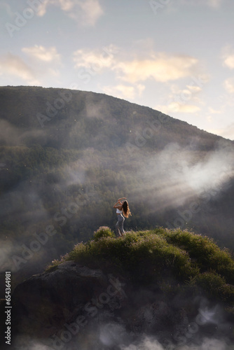 Beautiful young woman posing in the sunset rays of the sun while standing on a rock against the backdrop of forest and rocky mountains