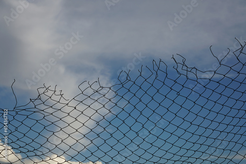 Looking up at a chain link fence with blue sky and clouds. wire fence. Chain link fence see sky. Opening in metallic fence. blue sky. Challenge. breakthrough concept. Chain-link  wire