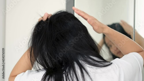 Asian woman wearing a white shirt looks in the mirror and touches her hair looking for gray hair or concerns about thinning hair