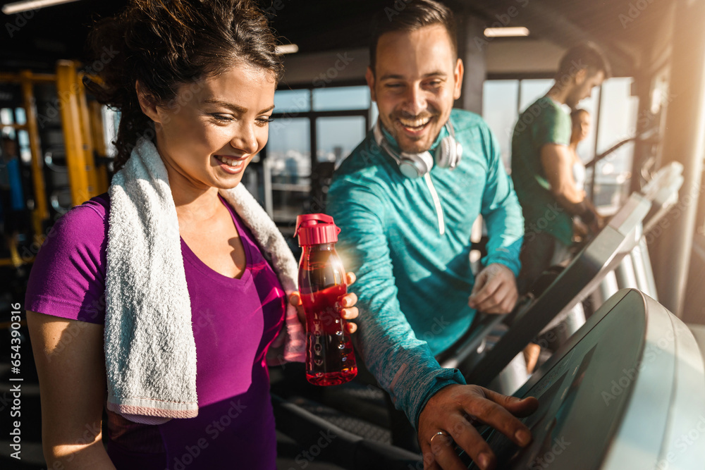 Happy athletic couple communicating while jogging on treadmills in a health club. Man explaining a woman how to turn on cardio machine. Attractive woman with water bottle needing assistance at gym. - obrazy, fototapety, plakaty 