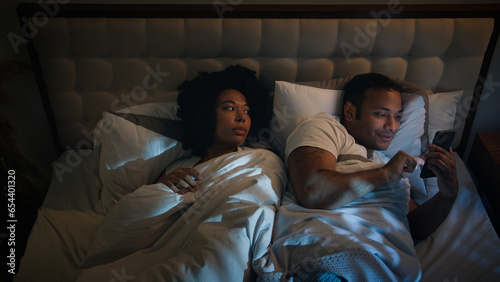 Irritated angry African American woman girlfriend trying sleep push man boyfriend husband using smartphone gadget addict addicted playing game online at late night mobile phone in bed couple family