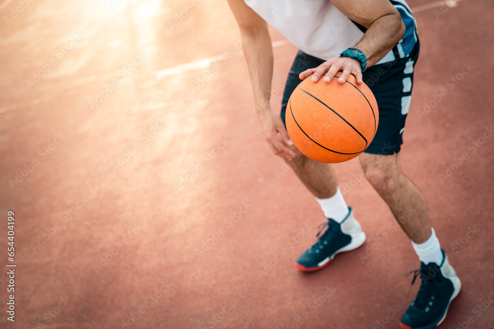 Sportsman playing basketball at basketball court outdoors. Photo of unrecognizable male person in movement dribbling a ball through playground. Focus on basketball.