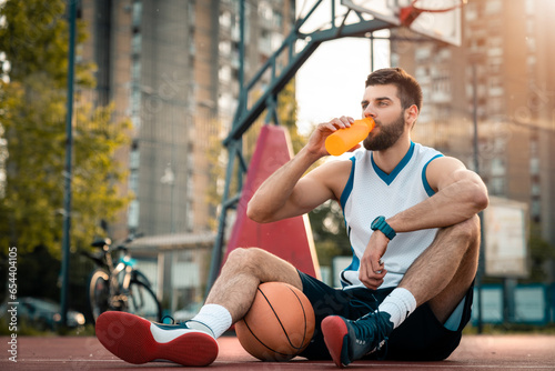 Tired basketball player drinking water from bottle while sitting at basketball court outdoors. Young man taking a break from sports activity rehydrating his body.