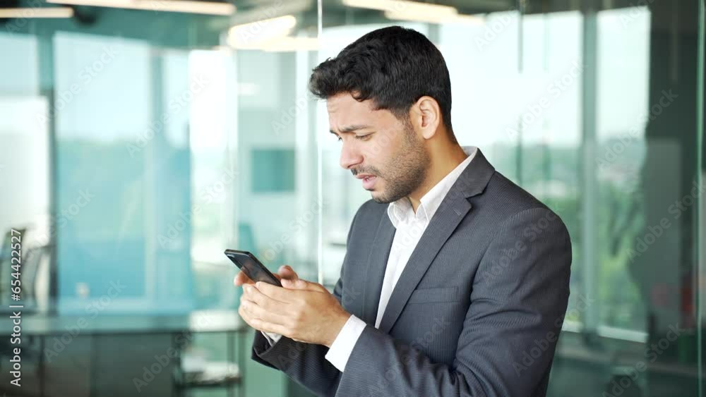 Side view. Frustrated stressed young businessman in a formal suit reads bad news on smartphone standing in business office. Disappointed entrepreneur is sad reviewing negative information on the phone Side view. Frustrated stressed young businessman in a formal suit reads bad news on smartphone standing in business office. Disappointed entrepreneur is sad reviewing negative information on the phone