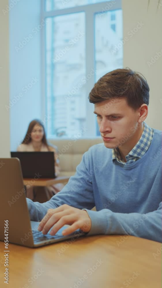 Vertical Screen: Serious man wearing sweater working on laptop in cafe, glass of water standing beside, woman with laptop on blurred background. Person browsing internet. Freelancer working remotely Vertical Screen: Serious man wearing sweater working on laptop in cafe, glass of water standing beside, woman with laptop on blurred background. Person browsing internet. Freelancer working remotely