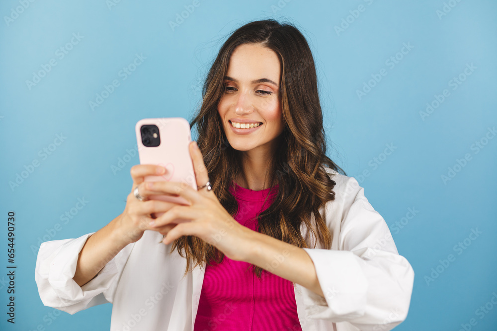 Happy carefree woman messaging. Smiling woman holding mobile phone device, doing commerce online shopping on cell, using apps on cellphone standing isolated on blue background, scrolling and typing. Happy carefree woman messaging. Smiling woman holding mobile phone device, doing commerce online shopping on cell, using apps on cellphone standing isolated on blue background, scrolling and typing.