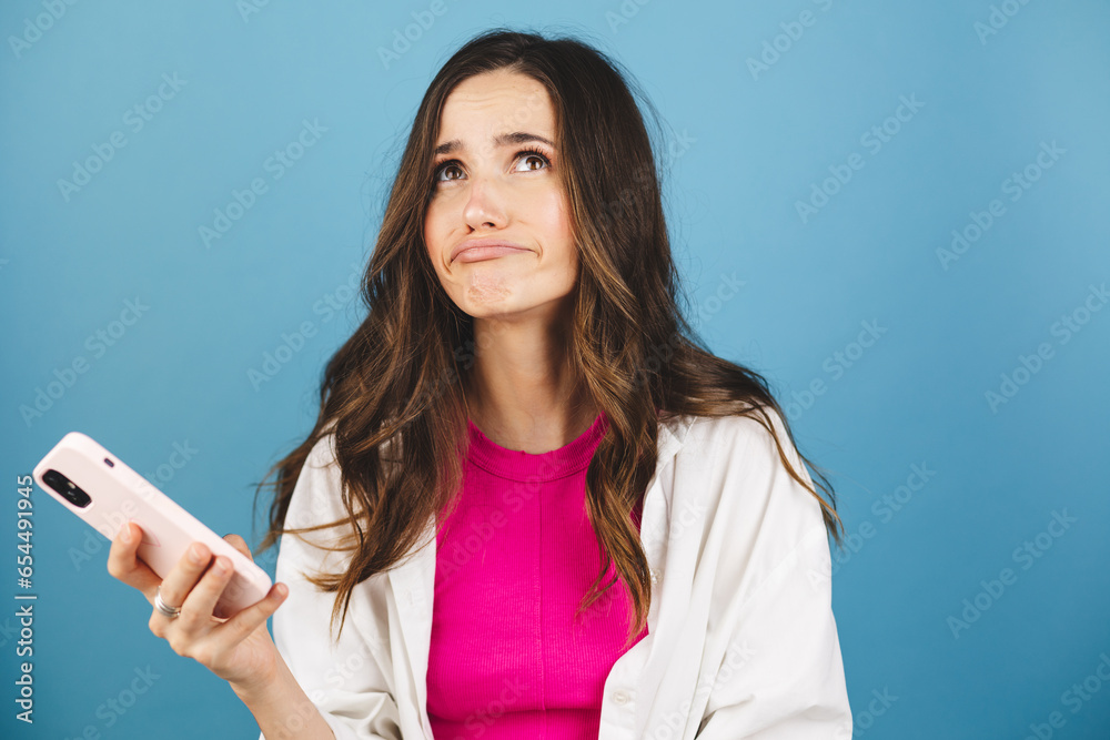 Brunette curly woman in pink top and white shirt looking sad disappointed girl holding in hands mobile phone reading bad negative news, gloomy lips isolated over bright blue color background. Brunette curly woman in pink top and white shirt looking sad disappointed girl holding in hands mobile phone reading bad negative news, gloomy lips isolated over bright blue color background.