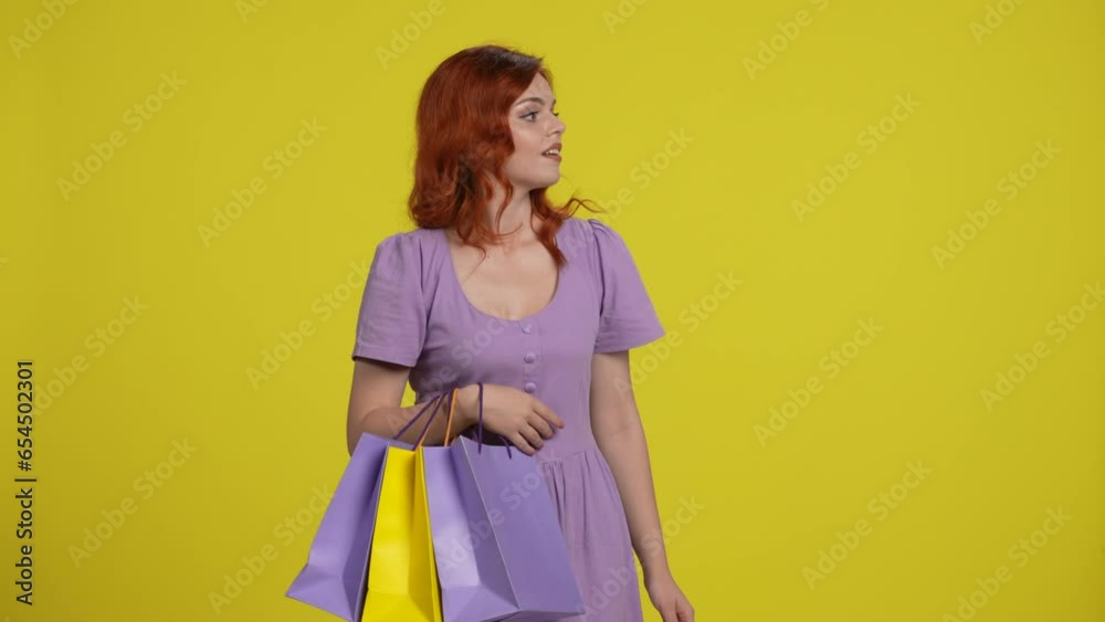 The redhaired woman looks around in search of someone, happily waves her hand in surprise as a sign of greeting. A woman pats shopping bags with her palm in a studio on a yellow background. The redhaired woman looks around in search of someone, happily waves her hand in surprise as a sign of greeting. A woman pats shopping bags with her palm in a studio on a yellow background.