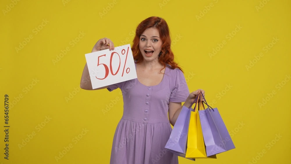A woman holds shopping bags and shows the inscription fifty percent. Satisfied shopaholic woman in studio on yellow background. A woman holds shopping bags and shows the inscription fifty percent. Satisfied shopaholic woman in studio on yellow background.