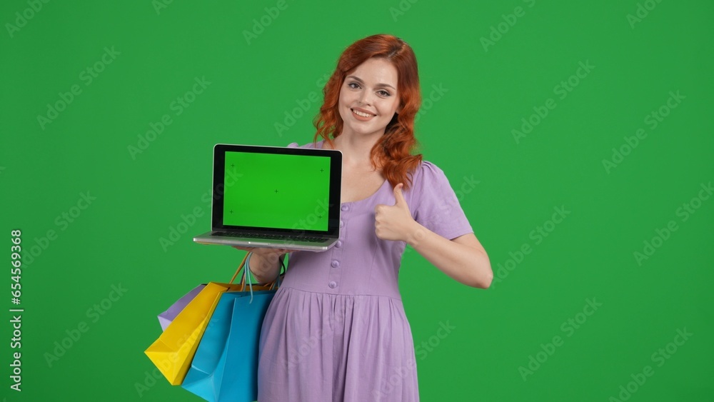 Young woman with shopping bags holding a laptop with green screen and showing thumbs up. Woman in studio on green screen. Advertising area, workspace. Sale, Black Friday, discounts. Young woman with shopping bags holding a laptop with green screen and showing thumbs up. Woman in studio on green screen. Advertising area, workspace. Sale, Black Friday, discounts.