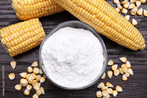 Bowl with corn starch, ripe cobs and kernels on dark wooden table, flat lay