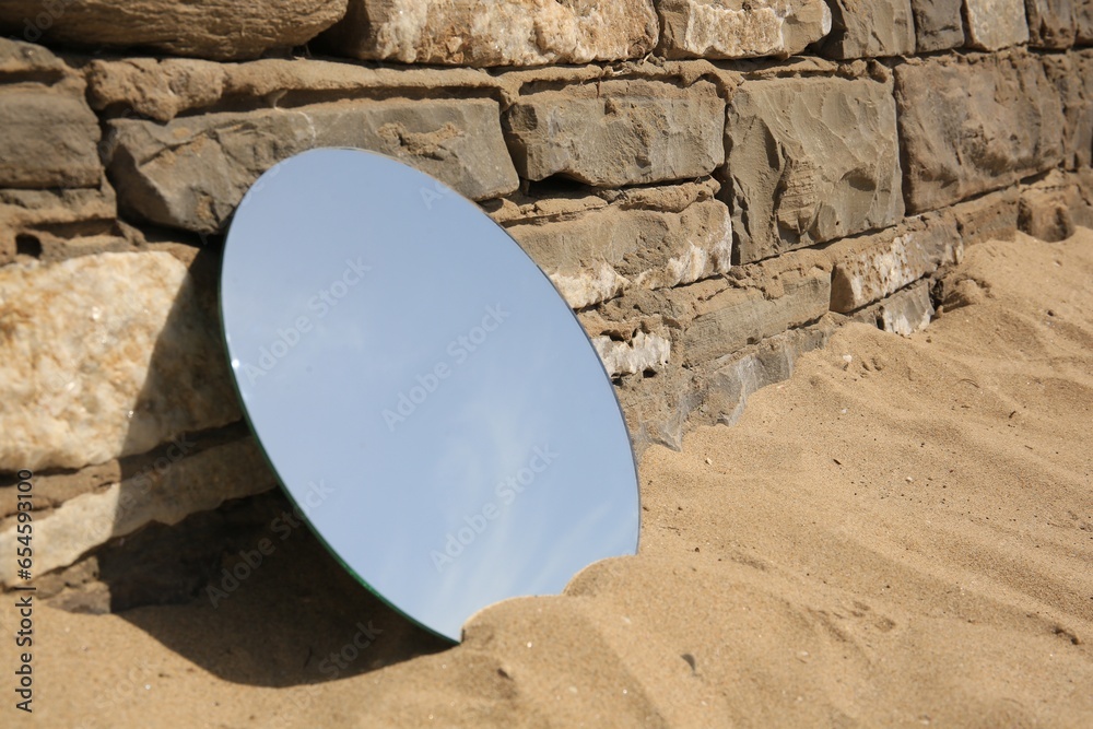 Round mirror reflecting sky on sand near stone wall outdoors, space for text