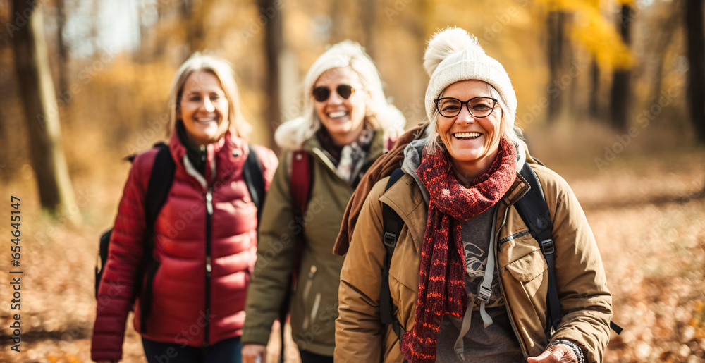 Positive, cheerful elderly women walk with backpacks through autumn forest park. Happy mature ladies travel in group