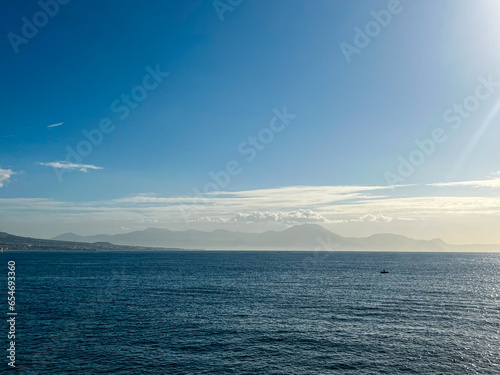 View at the mount Vesuvius from the opposite coast in Naples, Italy. Bright blue sky and fluffy clouds located around the volcano crater. The sea is calm. Small fishermen boats are at standstill
