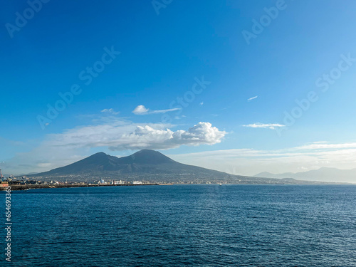 View at the mount Vesuvius from the opposite coast in Naples, Italy. Bright blue sky and fluffy clouds located around the volcano crater. The sea is calm. Small fishermen boats are at standstill
