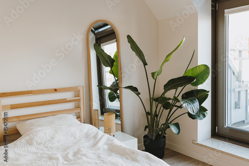 Modern Nordic Scandinavian home interior design. Bedroom with comfortable bed, tropical palm plant, mirror, lamp, wide window. Elegant apartment for rent concept photo