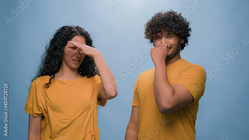 Bad smell reaction multiracial diverse couple Indian Arabian man and Hispanic woman cover noses suffer disgust stink stinky smelly aroma frowning sniffing unpleasant odor at blue studio background