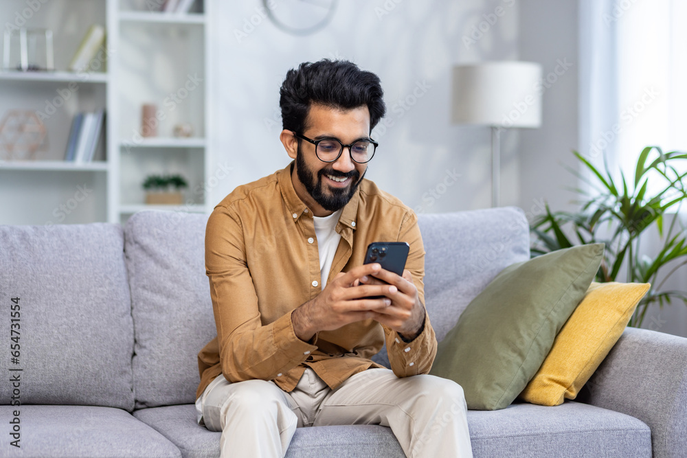 Young hispanic man sitting at home on sofa in living room of house, happy and smiling man holding phone, using smartphone app, typing message and watching video, online shopping